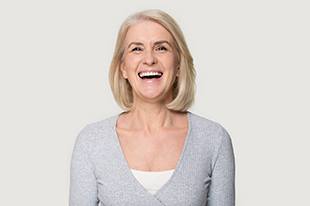 A middle-aged woman wearing a gray V-neck sweater and smiling wide to show off her new dental implants 
