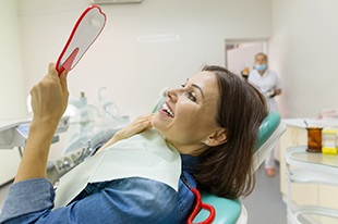A middle-aged woman with dark hair and wearing a denim shirt looking at her new smile in the mirror while at the dentist’s office 
